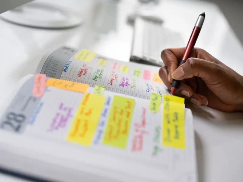 Calendar scheduling software being used as a person writes in a planner filled with colorful sticky notes